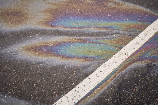 Colorful gasoline or oil spill creating a rainbow sheen on a dark asphalt road surface with a white traffic line.