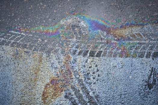 Spill of gasoline or oil with tire marks creates a rainbow sheen on the asphalt road surface.