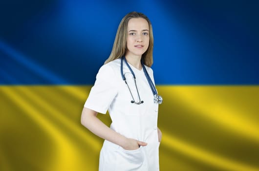 A young female doctor in a white coat and a stethoscope stands on the background of the flag of Ukraine.