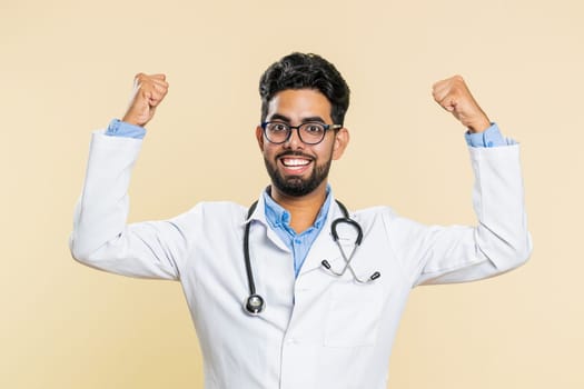 Indian young doctor cardiologist man celebrating success victory, winning, achievement, good positive news, triumph, succeed. Arabian apothecary pharmacy guy champion isolated on beige background