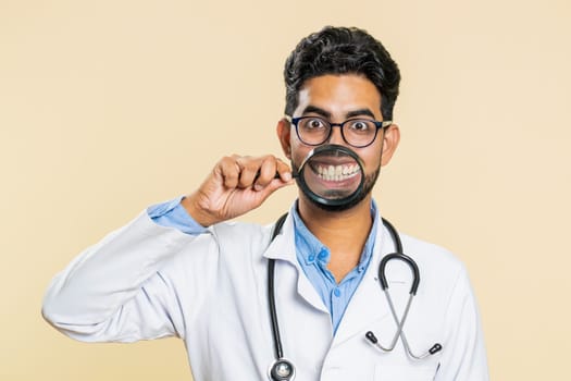 Happy Indian young doctor cardiologist man holding magnifier glass on teeth funny smiling silly face mouth. Health care, hygiene, stomatology. Arabian apothecary pharmacy guy on beige background