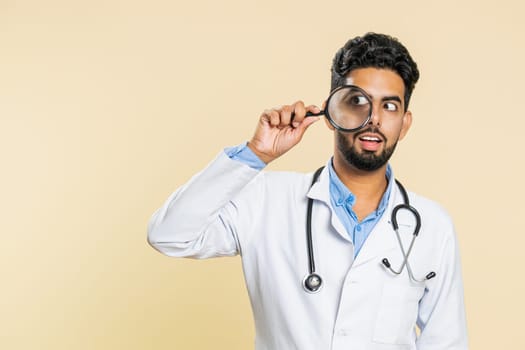 Investigator researcher scientist Indian young doctor cardiologist man holding magnifying glass near face looking , searching, analyzing. Arabian apothecary pharmacy guy isolated on beige background