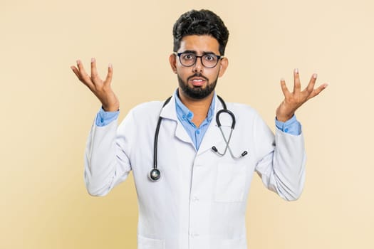 What. Why. Confused Indian doctor cardiologist man raising hands in indignant expression asking reason of failure, demonstrating disbelief irritation by troubles. Apothecary guy on beige background
