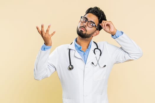 I don't know what to say. Confused Indian young doctor cardiologist man feeling embarrassed about ambiguous question, having doubts, no answer, clueless. Arabian apothecary guy on beige background