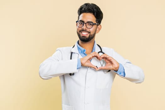 Indian doctor cardiologist man in love. Smiling attractive Arabian apothecary pharmacy guy makes heart gesture demonstrates love sign expresses good positive feelings isolated on beige background