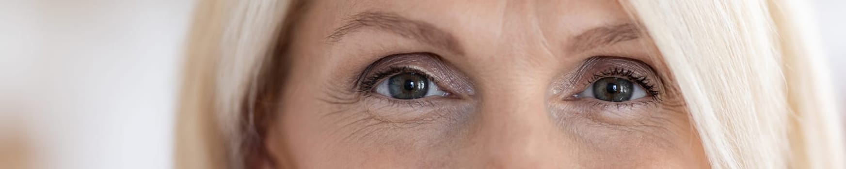 Eyes of attractive blonde senior woman in her 60s, blurred background, web-banner, panorama with copy space. Aesthetic medicine, plastic surgery, beauty treatment for elderly ladies