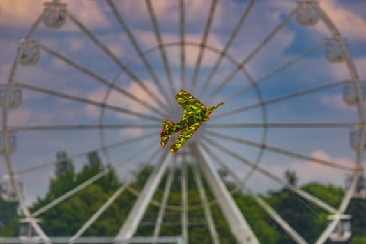 Small paper kite in shape of aeroplane flying in front of big white ferris wheel at cloudy afternoon