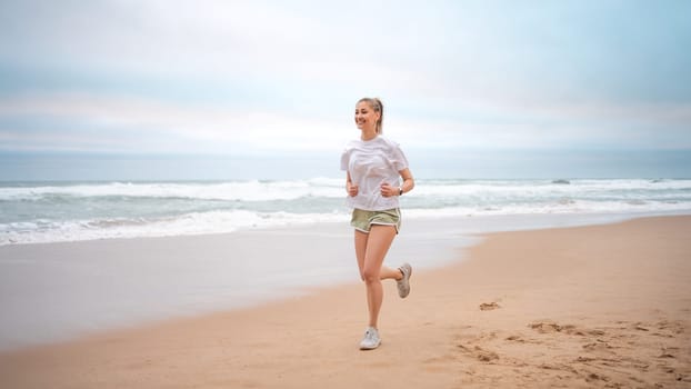 Woman jogging on beach. Sporty female jogging on sand beach, waves on background. Full body view woman in mini shorts and t-shirt who runs alongside ocean. Outdoor workout of trained fit girl.