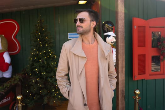 Fashionable man stands next to Christmas tree. Festive location: good looking man in beige coat enjoying Christmas city decorations. Elegant male wearing sunglasses watching Christmas city attractions