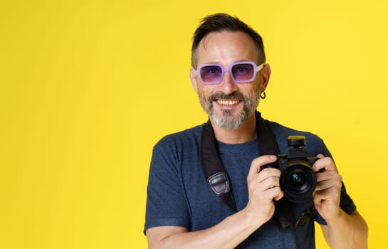 Happy, enthusiastic photographer smiles with delight while holding mirrorless camera against yellow background. Joy and passion that photographer feels when capturing moments with beloved camera. High quality photo