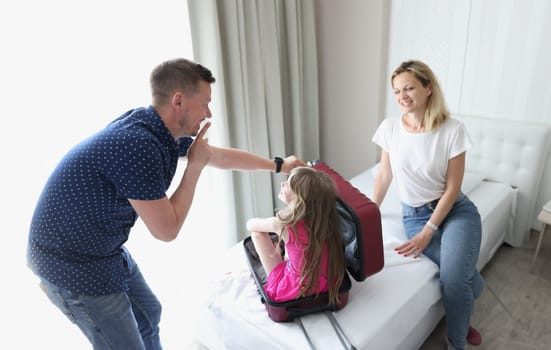 Cheerful dad and mom play hide and seek with child and suitcase. Dad asks child to sit quietly in suitcase