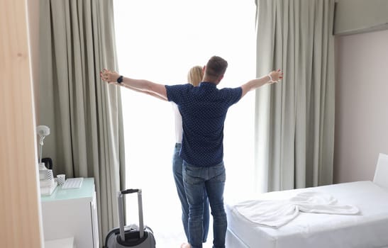 Married couple hugging and holding hands at window of hotel room in bedroom. Love romantic travel and honeymoon