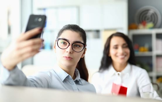 Young woman in glasses holds phone in front of face and smiles at colleagues in office. Video call or selfie at work