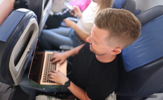 Male freelancer is busy working with laptop while sitting on airplane during business trip. Remote work or watching movies information on Internet in airplane