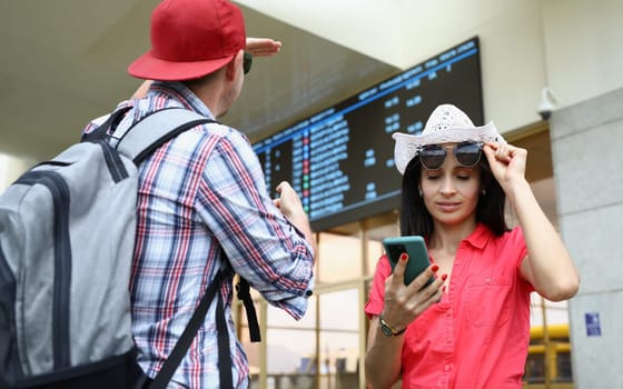 Man and woman are tourists with mobile phone against background of electronic board of bus and railway transport timetables. Smartphone airport transport concept app and flight schedules