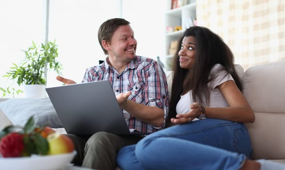 Interethnic family couple laughing and chatting while sitting on sofa at home with laptop. Married couple making online payments on laptop using financial banking app on computer at home