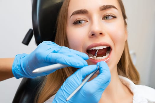 Woman patient sitting on chair during teeth procedures in dental clinic. Dentist checks front teeth with help of mirror tool and checks strength of teeth