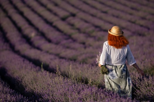 Here's a caption under 150 characters describing the image: Redhead woman in straw hat walks through a vast, purple lavender field. Rows of lavender stretch to the horizon.