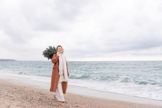 Blond woman Christmas sea. Christmas portrait of a happy woman walking along the beach and holding a Christmas tree on her shoulder. She is wearing a brown coat and a white suit