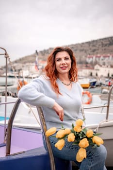 Woman holds yellow tulips in harbor with boats docked in the background., overcast day, yellow sweater, mountains.