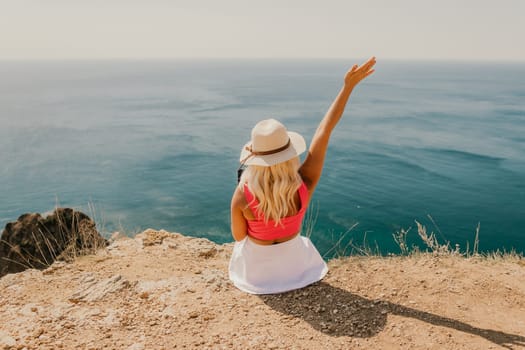 A woman is sitting on a rocky beach with a laptop in front of her. She is wearing a pink top and a straw hat. She is waving her hand in the air, possibly to celebrate or to show excitement
