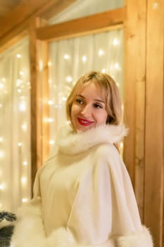 A blonde woman in a white fur coat smiles and looks over her shoulder, standing in front of a string of lights.