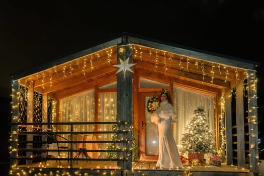 A woman in a white dress stands on a porch, illuminated by string lights, in front of a Christmas tree inside a wooden cabin.
