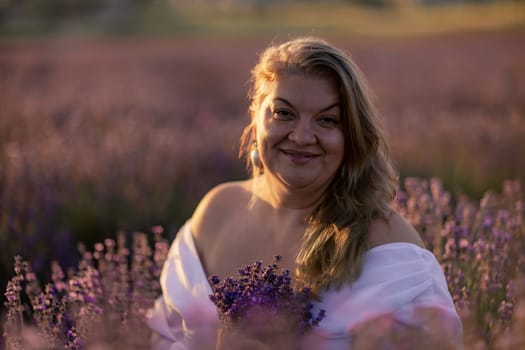 Woman poses in lavender field at sunset. Happy woman in white dress holds lavender bouquet. Aromatherapy concept, lavender oil, photo session in lavender.