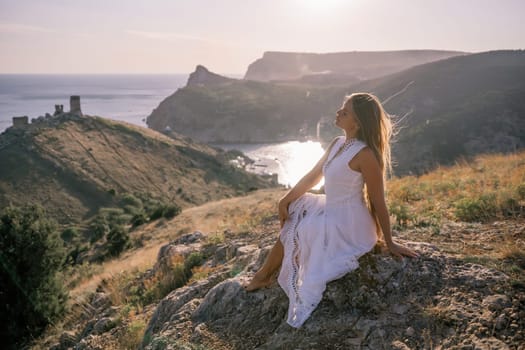A woman in a white dress is sitting on a rock overlooking a body of water. She is enjoying the view and taking in the scenery