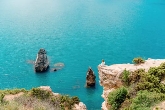 A woman sits on a rock overlooking the ocean. The water is blue and calm. The scene is peaceful and serene