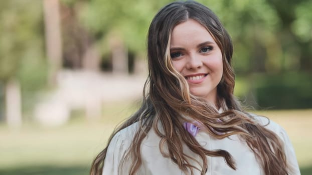 Portrait of a cheerful young woman with long, flowing hair, enjoying a sunny day in a park, wearing a stylish white shirt