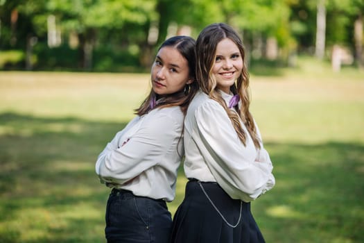Two cheerful students pose with crossed arms, back to back in a park, beaming on their first day of school