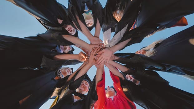 Group of happy successful graduates in academic hats and robes standing in circle and putting their hands together. Team of college or university students celebrating graduation.