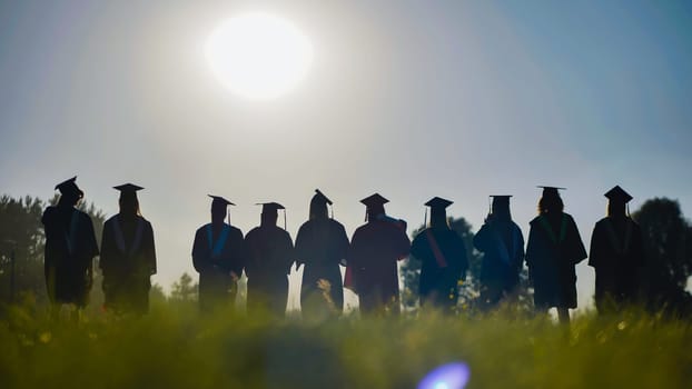 Silhouetted graduates wearing gowns and caps stand in a field at sunset, celebrating their high school commencement