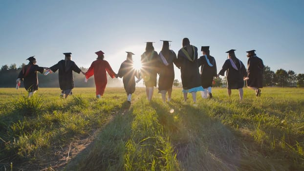 Group of students wearing graduation gowns walking away in a field at sunset, holding hands and celebrating their achievement