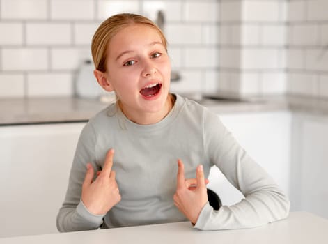 Cute Little Girl Sitting In The Kitchen At Home And Pointing Fingers At Herself