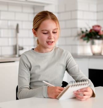 Happy Cute Little Girl At Home Writing An Essay In Notebook