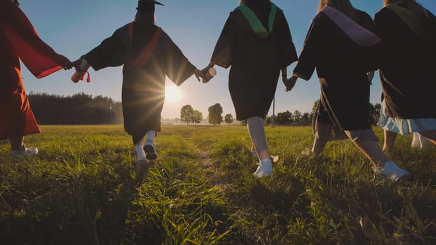 Group of cheerful graduates running together in a field at sunset, holding hands and celebrating their academic achievement