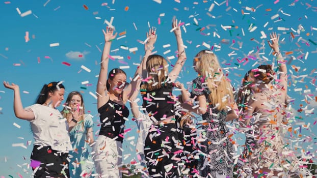 Group of teenage girls throwing colorful confetti in the air, celebrating graduation under a blue sky