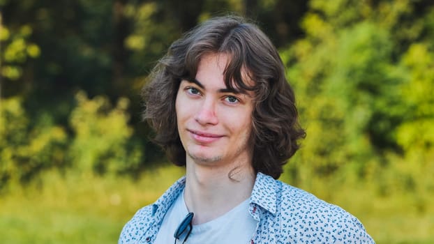 Young man with long brown hair is smiling while posing outdoors with a forest in the background
