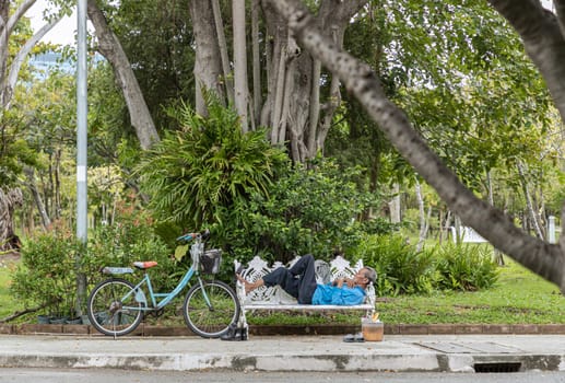 Bangkok, Thailand - 17 Oct, 2024 - View of An old man uses a lying on public bench to take an afternoon nap alongside old bicycle parked in a shady area of the quiet city park area. Elderly old senior man taking sleeping on bench, Space for text, Selective focus.