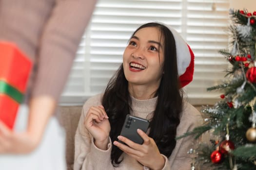 A young woman couple shares a joyful moment exchanging a surprise gift box on Christmas Day, surrounded by festive decorations.