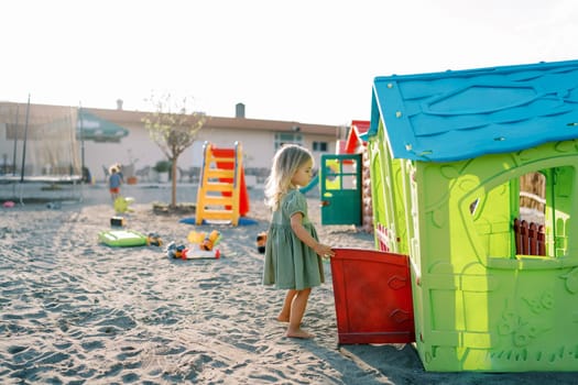 Little girl stands near a toy house with the door open and looking inside. High quality photo