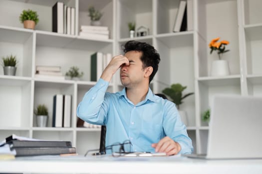 An office worker in a blue shirt struggles with a headache, feeling exhausted from intense work in a contemporary office setting.