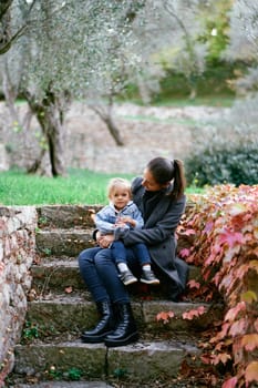 Mom with a little girl with a toy in her hands sits on stone steps in the park and looks at her. High quality photo