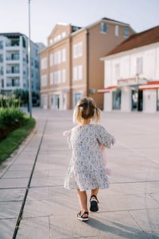 Little girl with a pink toy rabbit walks along the road to an apartment building. Back view. High quality photo