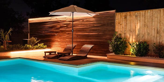 Modern illuminated swimming pool with wooden lounge chairs and a cozy umbrella creating a peaceful backyard ambiance. Concept of outdoor comfort and luxury at night