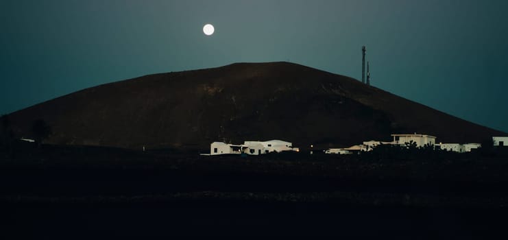 A road and a town with cozy white houses at dusk, illuminated by the light of the full moon against the backdrop of dramatic black land, volcanic origin and mountains of the Canary island of Lanzarote