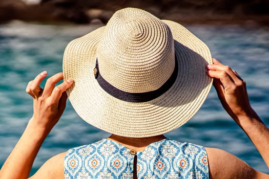 A young lady in a beautiful dress with ornaments holds a stylish hat in her hands and enjoys a leisure time, a vacation at sea. The girl stands with her back and looks at the ocean on a sunny day.