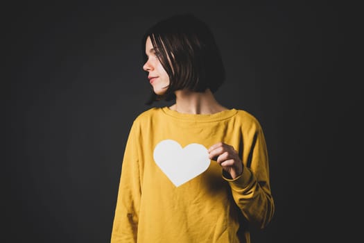 A young girl holds a white paper heart - a postcard or shows feelings of love. A woman with a joyful surprised face before the holiday and in search of love, relationships or shares her impressions.